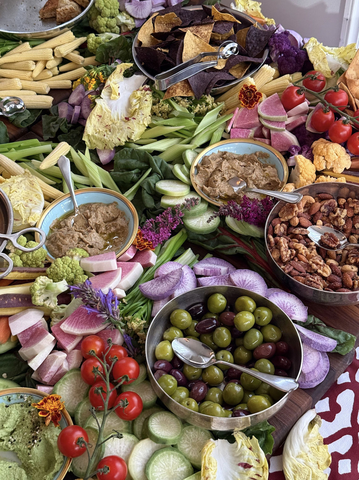 Colorful grazing table with fresh vegetables, dips, olives, nuts, tortilla chips, and edible flowers.
